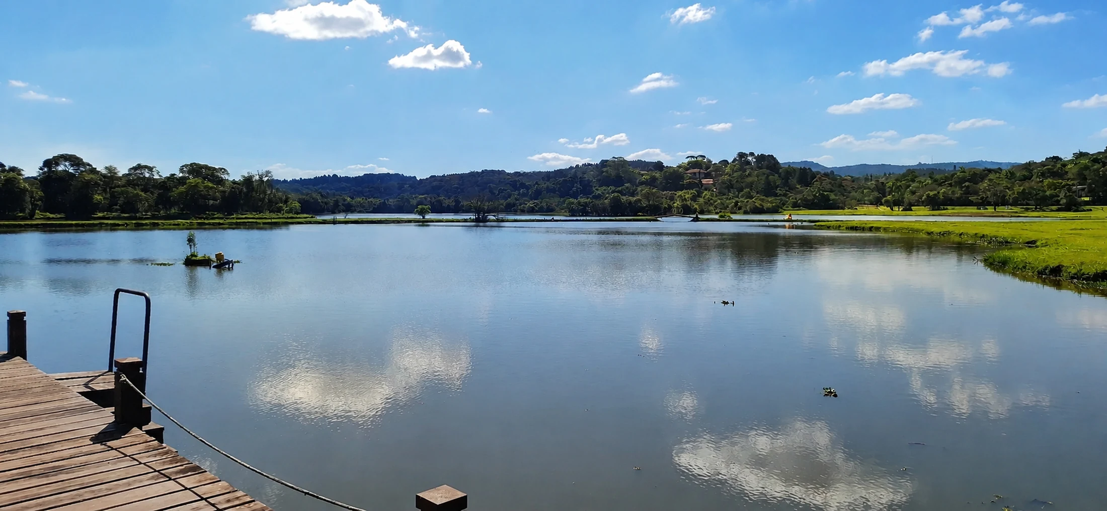 Lago do Patrimônio do Carmo com a mata refletida na água e um deck de madeira em primeiro plano